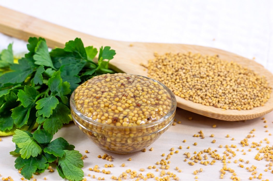 Yellow Dijon mustard sauce in glass dip with wooden spoon full of dry mustard seeds on the table in the kitchen.